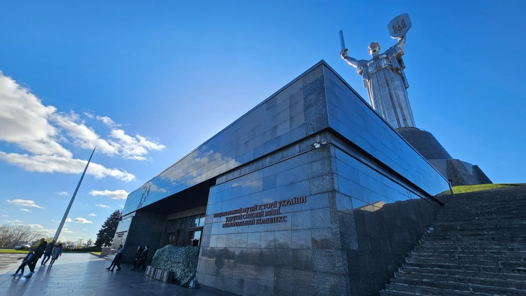 Entrance of the museum of the history of Ukraine in the Second World War in the foreground. In front of it, some sandbags and ammunition boxes. Some visitors are seen exiting the building. In the background, statue of Mother Ukraine. 