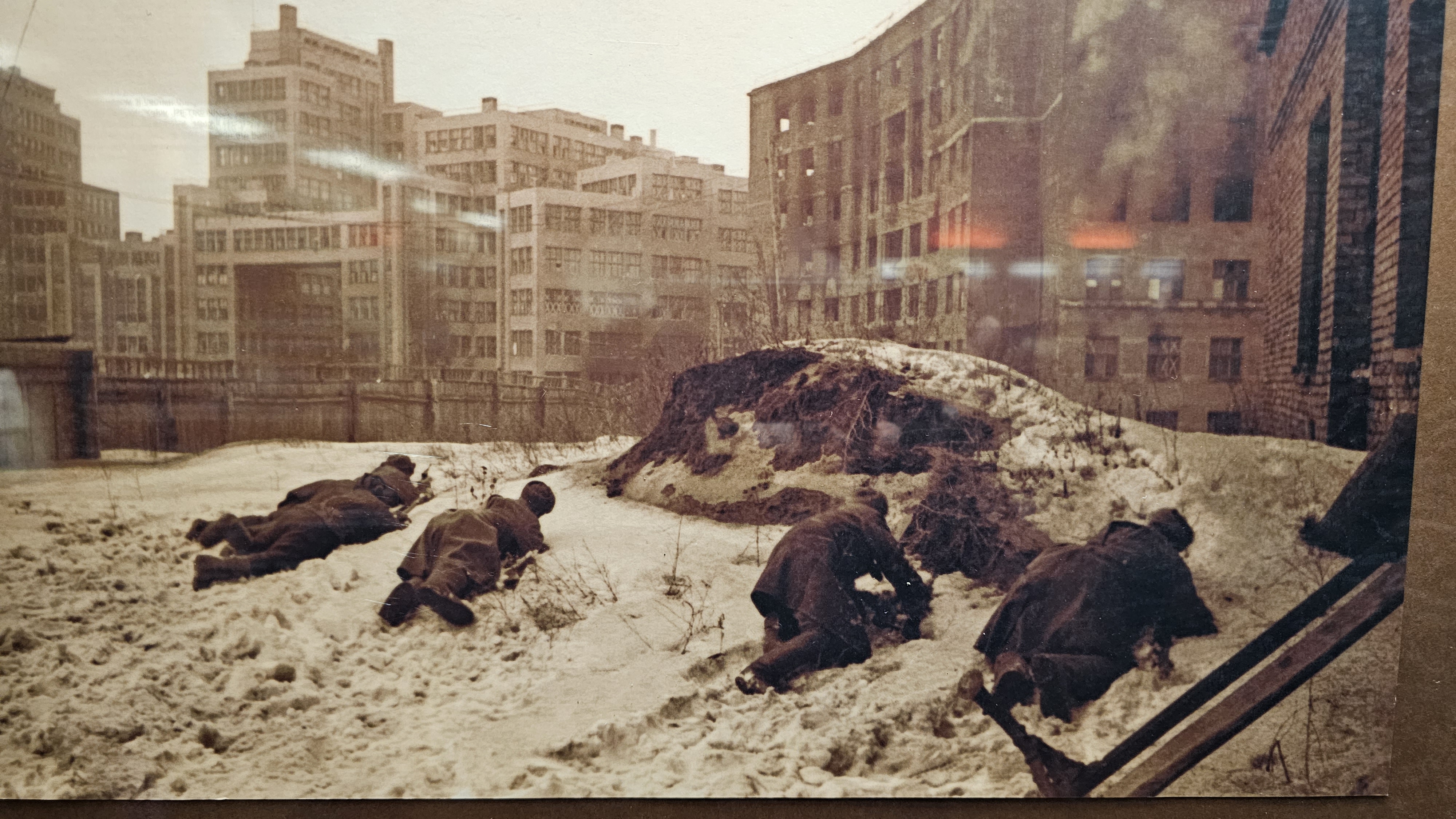 A sepia toned picture of soldiers lying in the snow in Kharkiv's city center. In the background, smoke is coming out of a destroyed building; Landmarks of the city are recognizable (today's main square).
