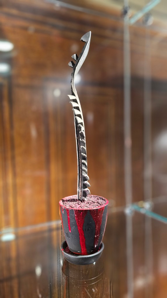 A dented aluminum fragment of missile is planted vertically into a base, looking like a metallic houseplant. Red paint covers the base.