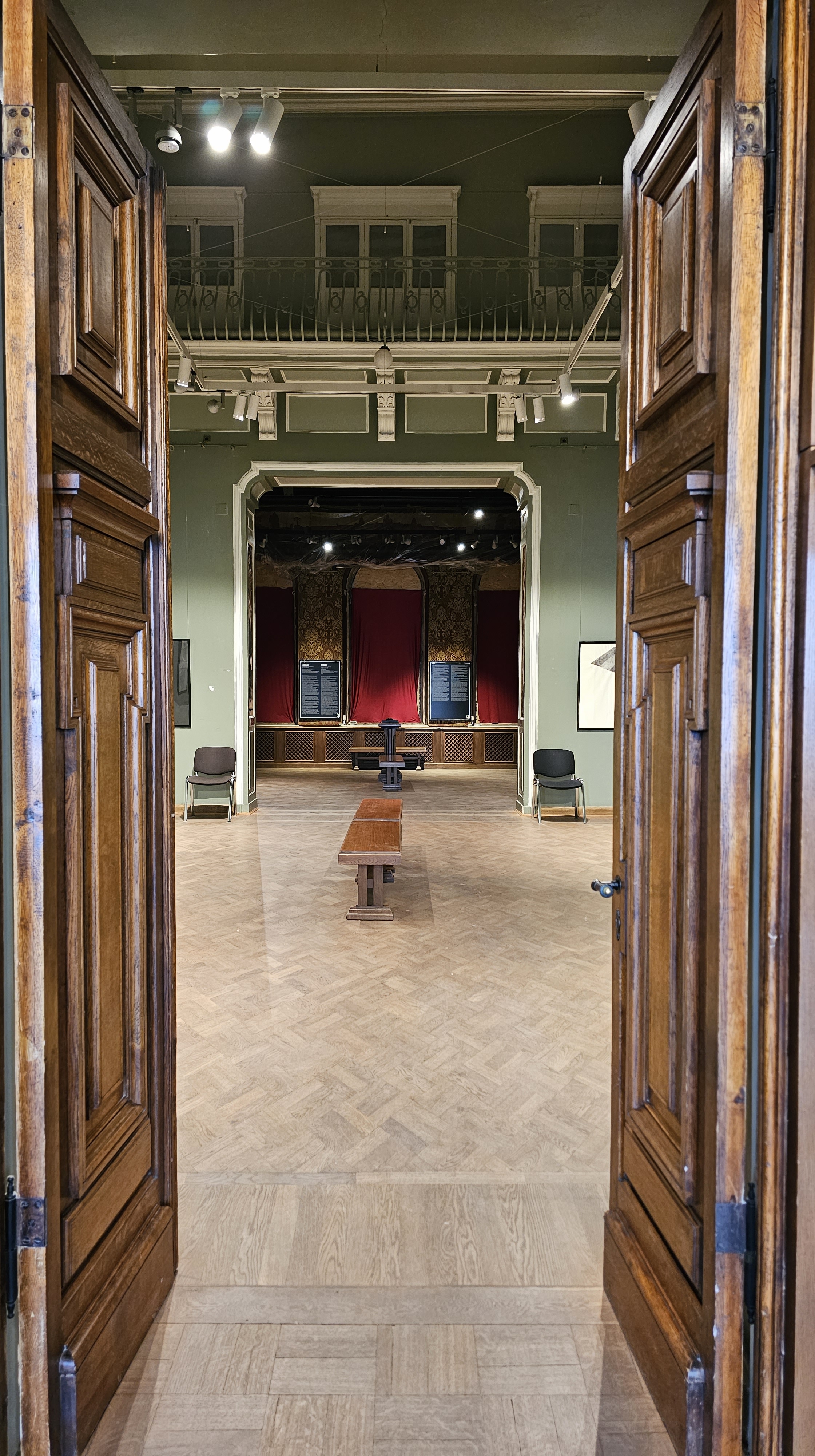 Two wooden doors open on an exhibition room with almond green walls and a wooden bench in th middle.