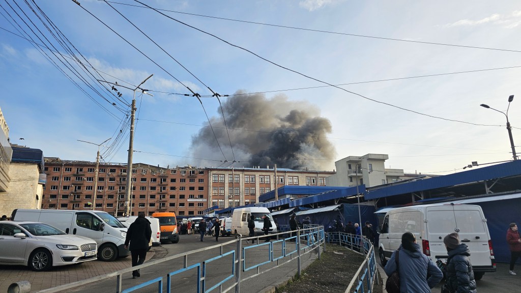 A dark cloud of smoke rises from the site of a missile strike near the subway station of Lukianivska, in Kyiv. People look on.