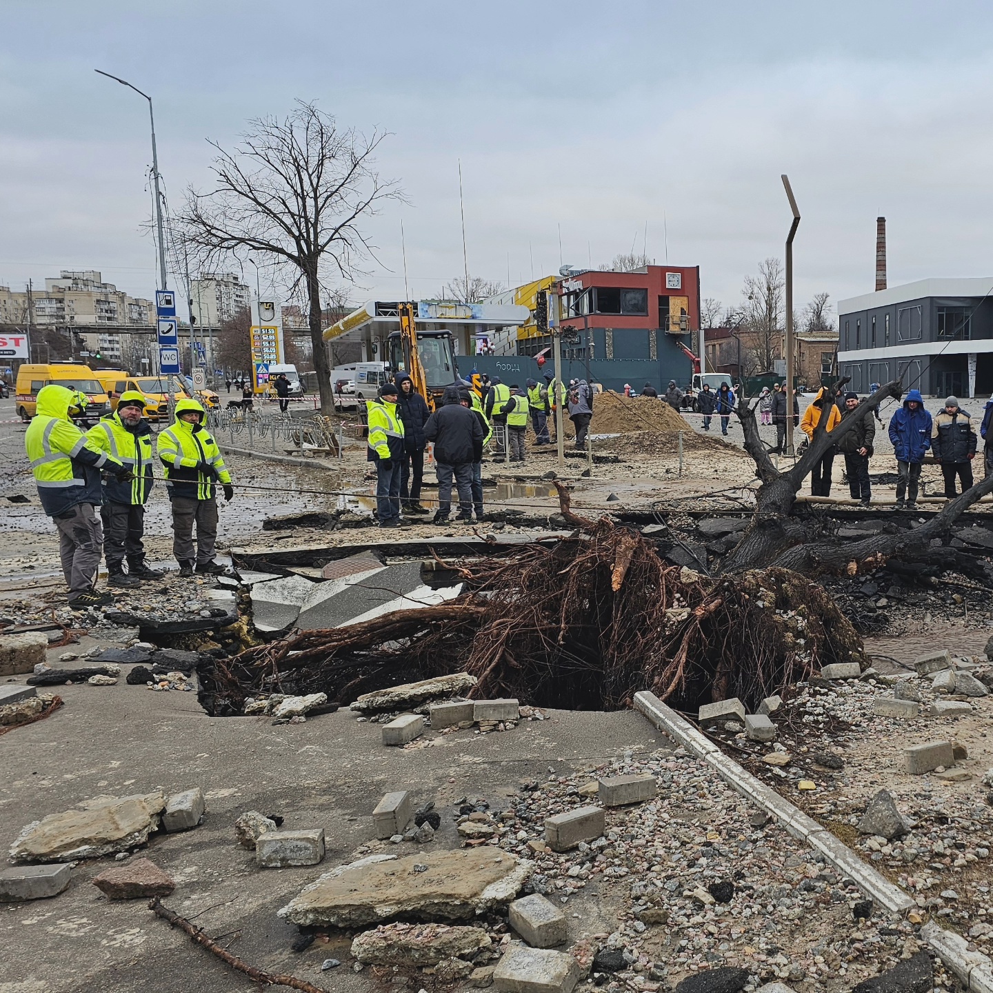Emergency workers secure the site of a missile strike in Podil district, they're around the crater left by the missile, in which a charred tree lies. Electric cables lie around.