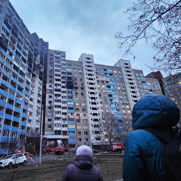 On the foreground, two people, a woman dressed with a purple jacket and a pink woollen hat, and a man in a blue hooded jacket, look on. In th background, a 20-stories residential building is partially destroyed by a strike, there are huge traces of smoke and dozens of broken windows.