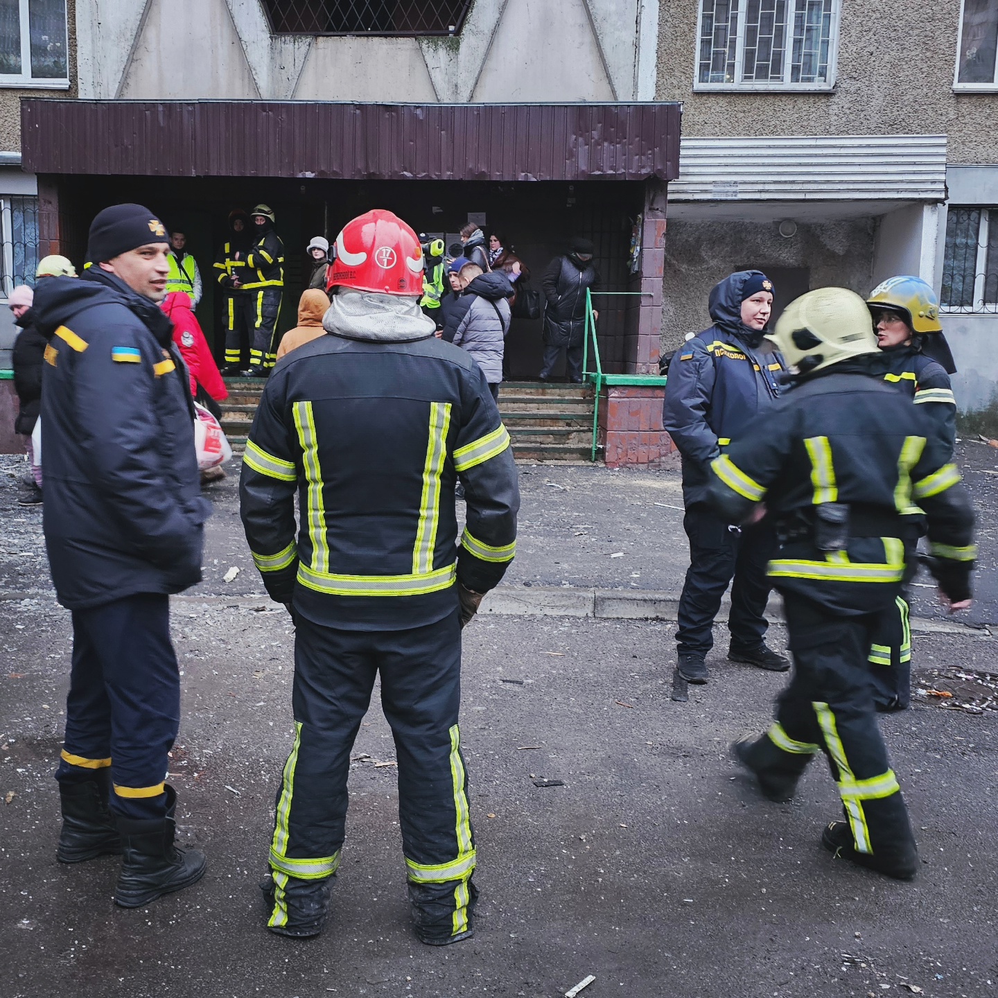 Firefighters are tending to residents waiting to retrieve their belongings next to a building hit by a Russian missile. There are glass shards on the ground.