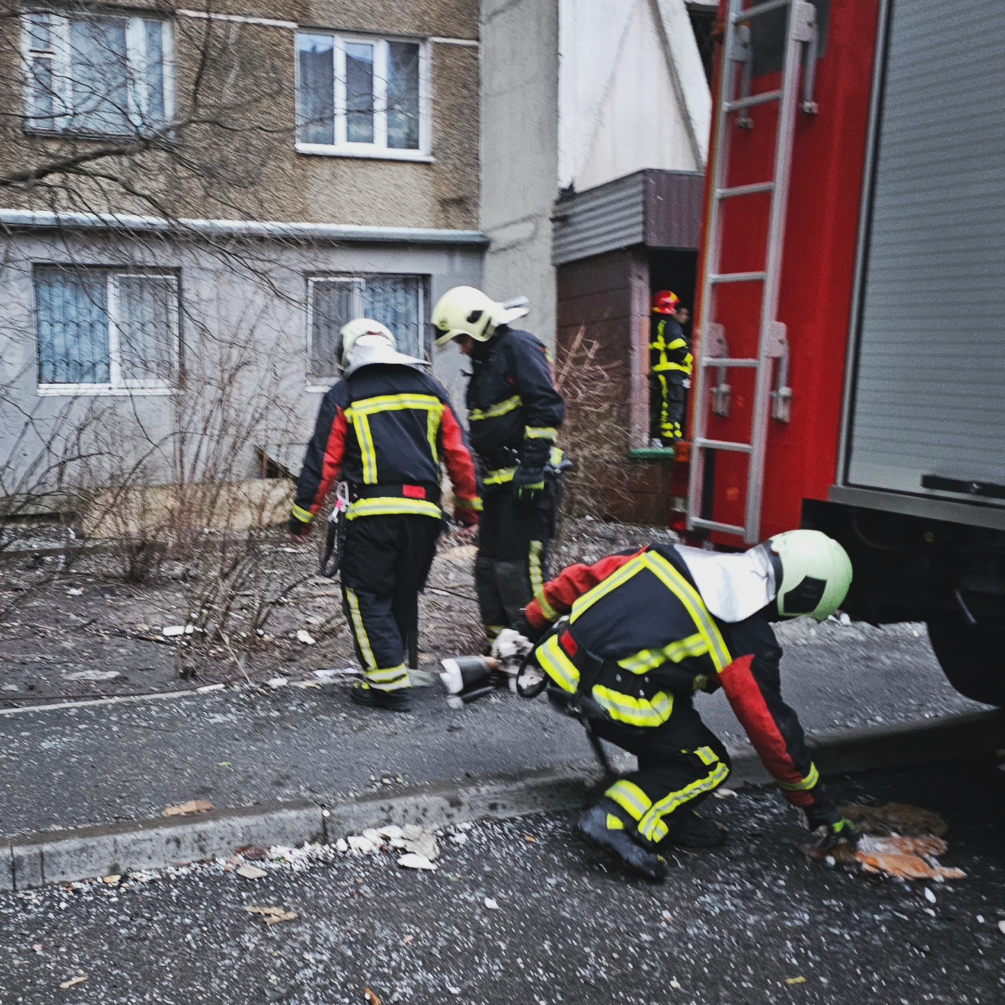 Three firefighters are clearing-up the pavement next to a building hit by a Russian missile. There are glass shards on the ground.