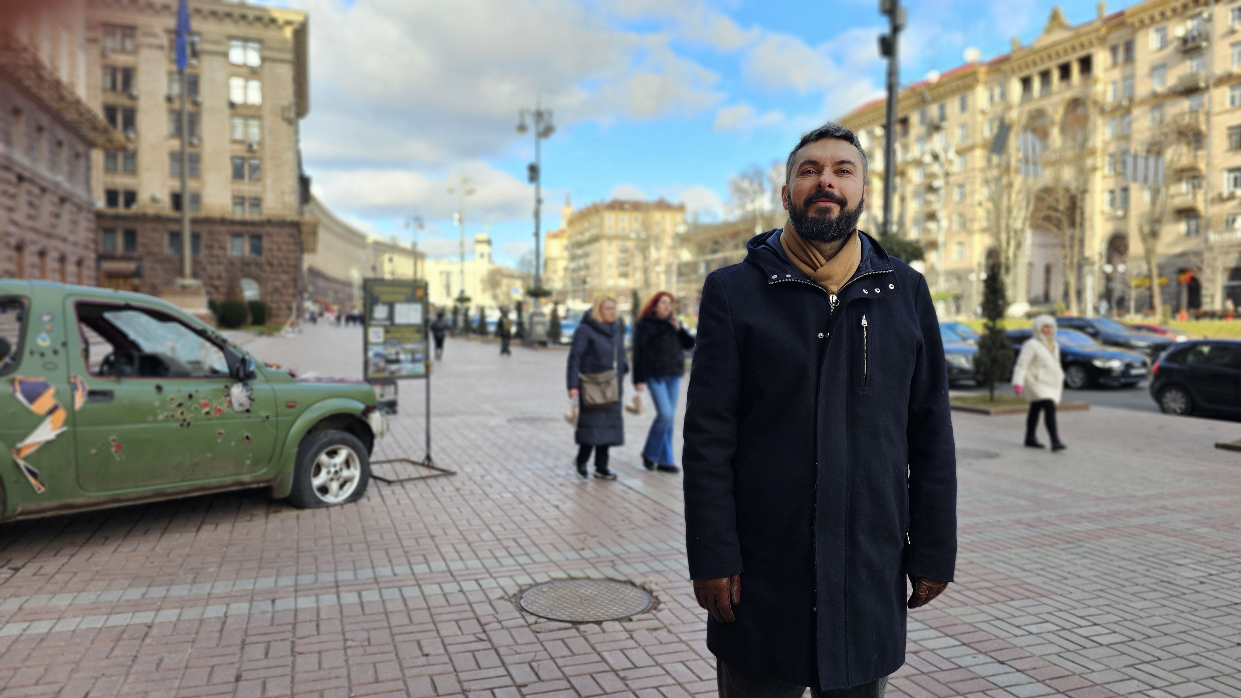 Max Kolesnikov, a year after his liberation, on Khreshchatik in Central Kyiv.
Behind him, a bullet-ridden civilian vehicle. 