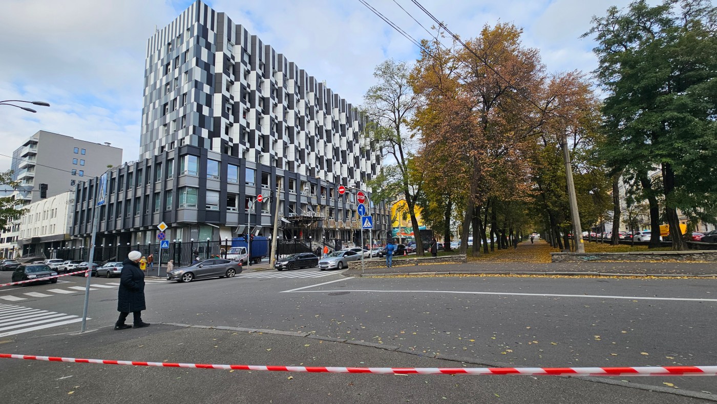 A police red and white cordon in the foreground. In the background, a street in Podil and one of the buildings impacted by a recent russian drones strike. the facade is partially destroyed, a lot of windows are shattered. People are working on the scene.